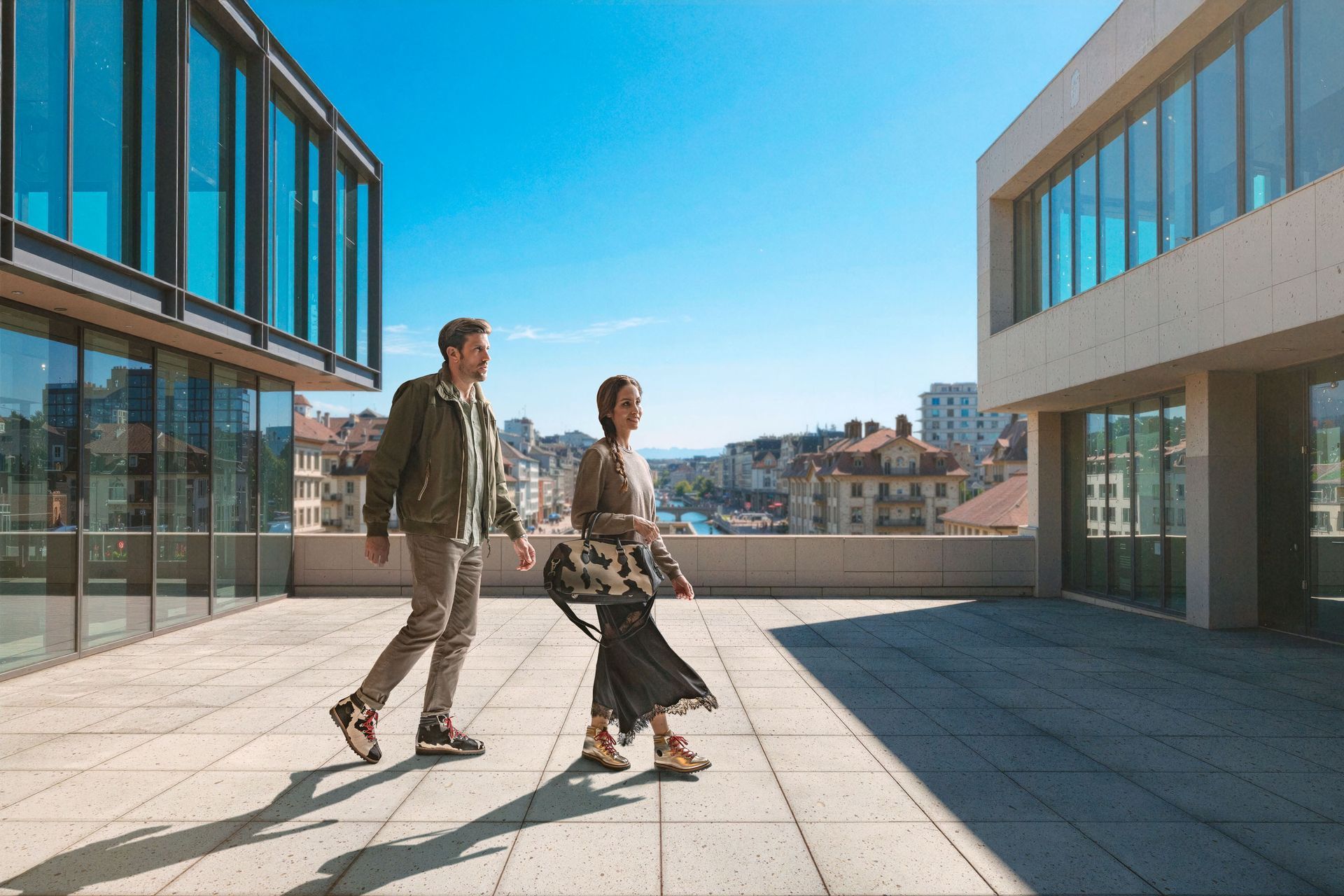 Zwei Personen gehen auf einer sonnigen Außenterrasse in der Nähe moderner Gebäude spazieren.