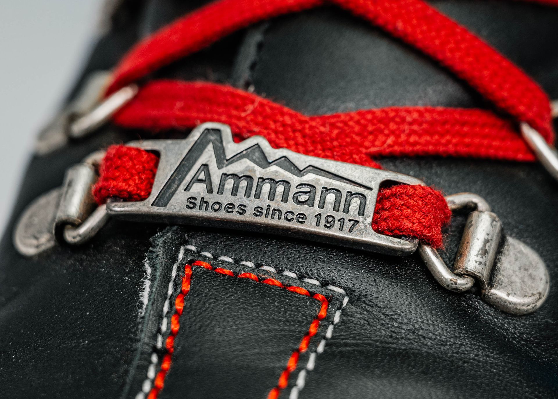 Schwarzes Stiefeldetail mit roten Schnürsenkeln, Ammann-Etikett aus Metall, weiße Nähte, Aufschrift „Schuhe seit 1917“.