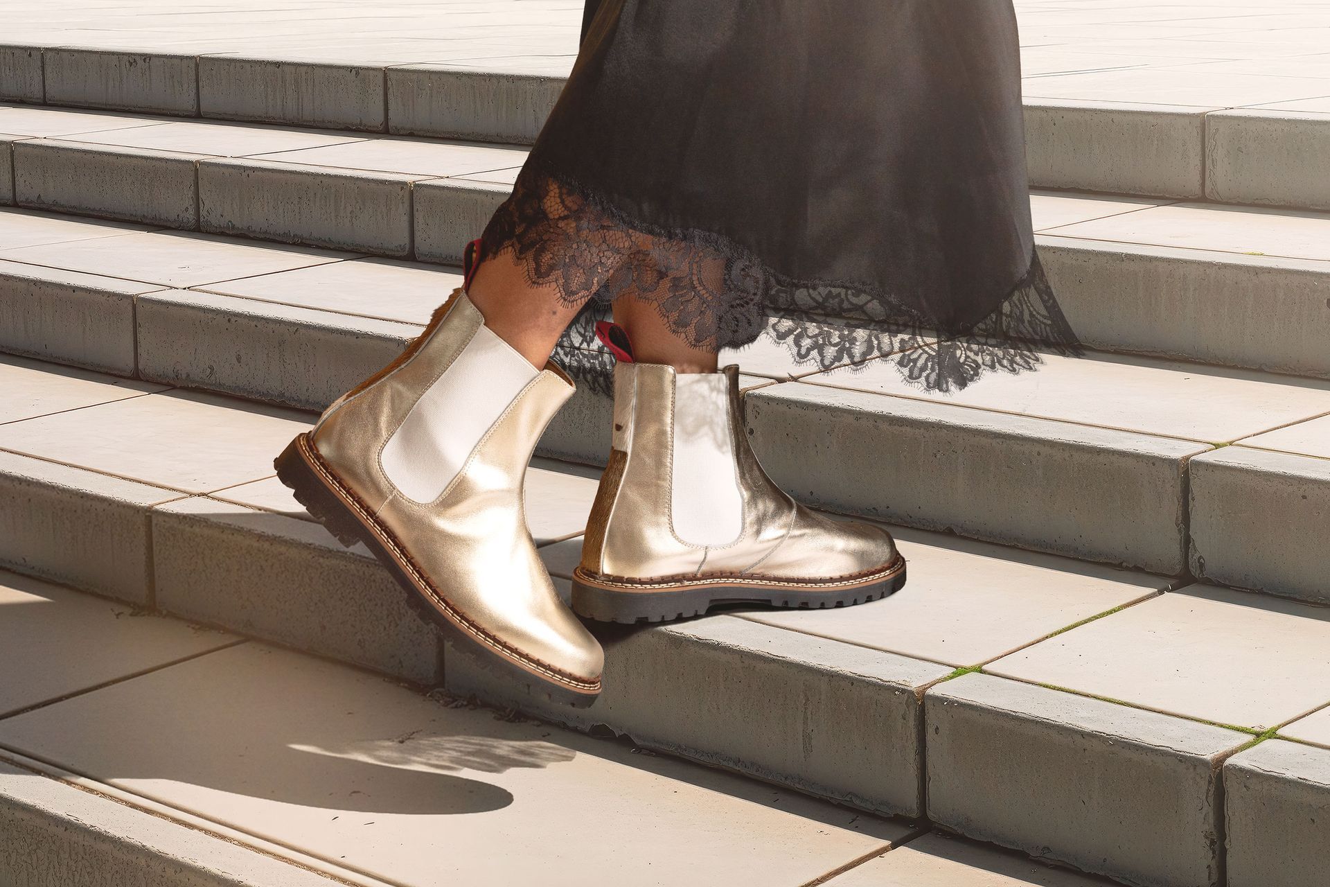 Eine Person trägt goldene Chelsea-Stiefel, die eine Betontreppe im Freien hinabsteigen; ein schwarzer Rock mit Spitzenbesatz.