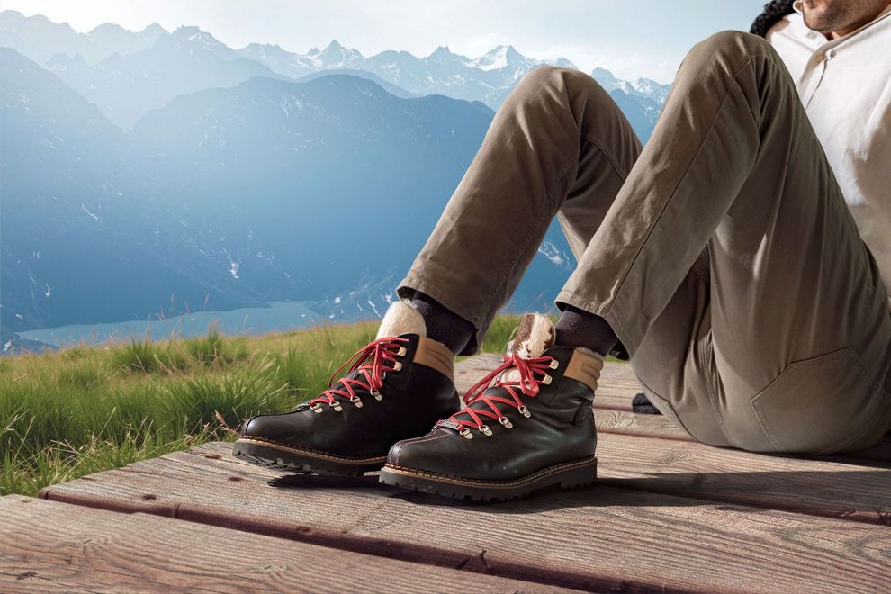 Person sitzt auf Holzoberfläche, trägt Wanderschuhe mit roten Schnürsenkeln, Bergkulisse.
