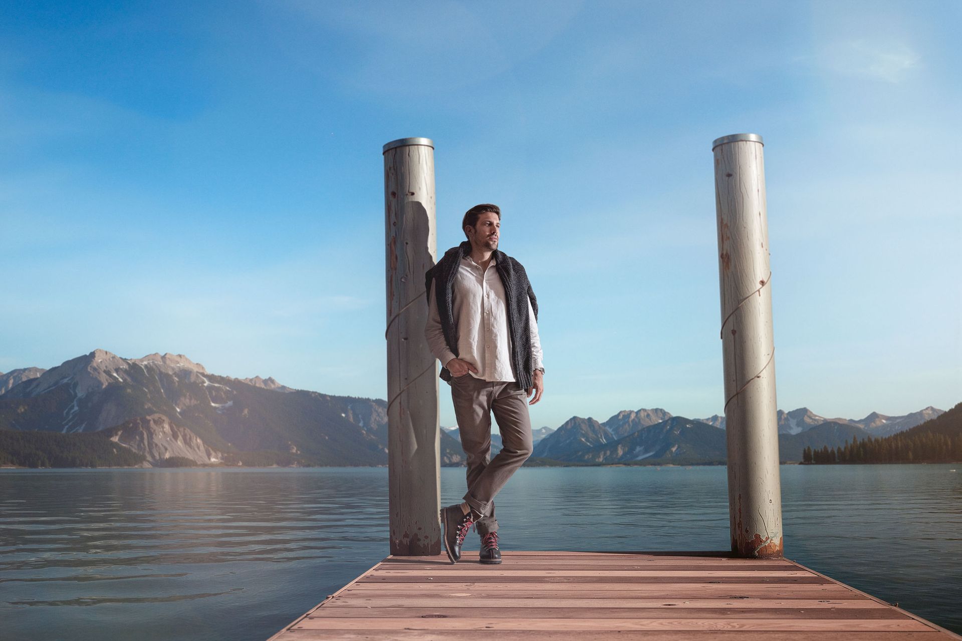 Mann lehnt an Dockpfosten an einem See, Berge im Hintergrund, sonniger Tag.