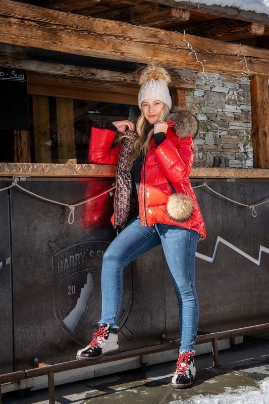 Frau in roter Pufferjacke und Jeans posiert in einer Bar im Freien in einer verschneiten Umgebung.