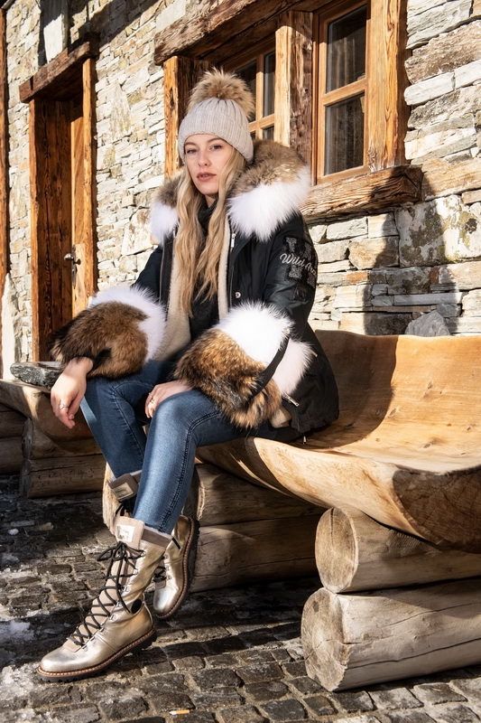 Frau im Wintermantel mit Pelzbesatz, auf einer Holzbank sitzend, mit Mütze und Stiefeln bekleidet.