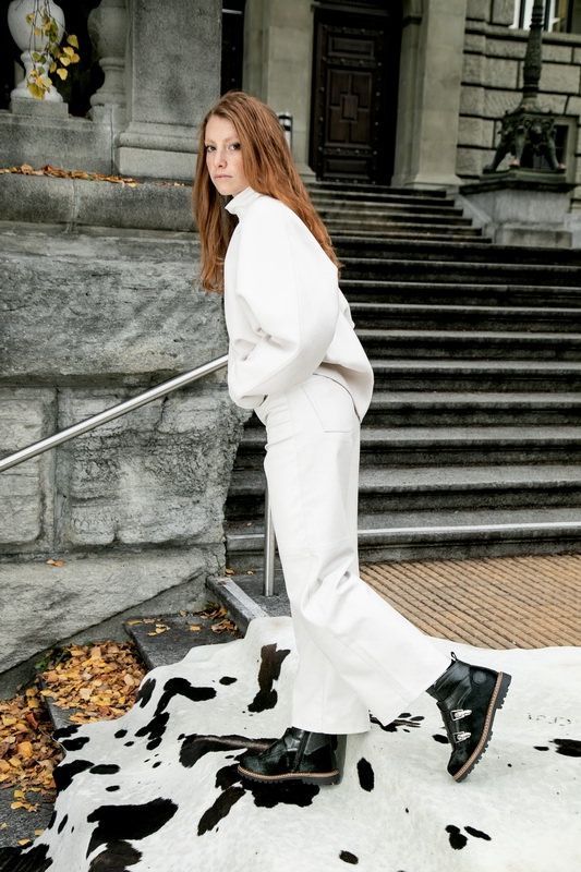 Frau in weißem Outfit und schwarzen Stiefeln geht auf einem Kuhfellteppich eine Außentreppe hinauf.