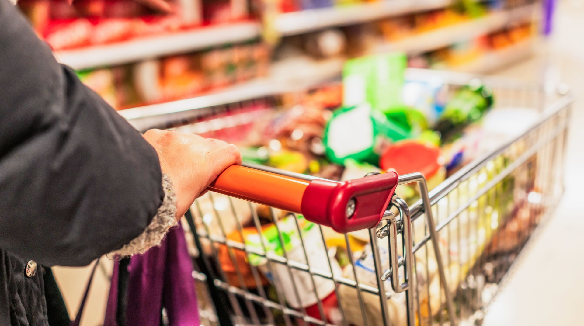 Persona che spinge un carrello della spesa pieno lungo il corridoio di un supermercato.