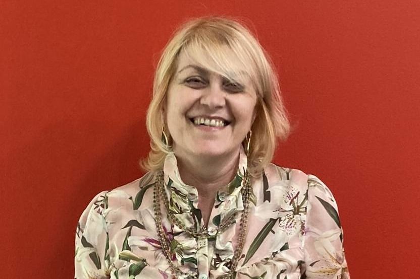 Donna dai capelli biondi che sorride davanti a un muro rosso, indossando una camicetta a fiori.