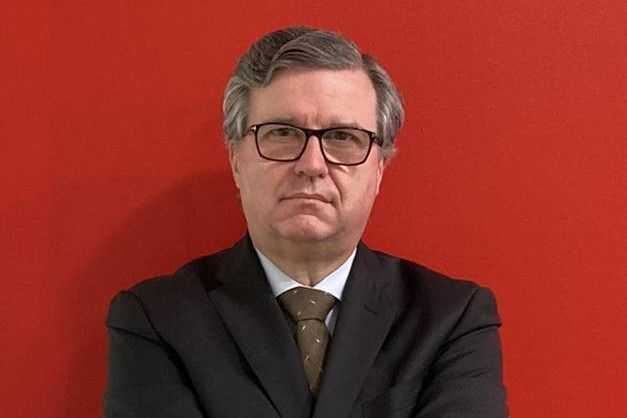 Uomo con occhiali e giacca, espressione seria, su sfondo rosso.