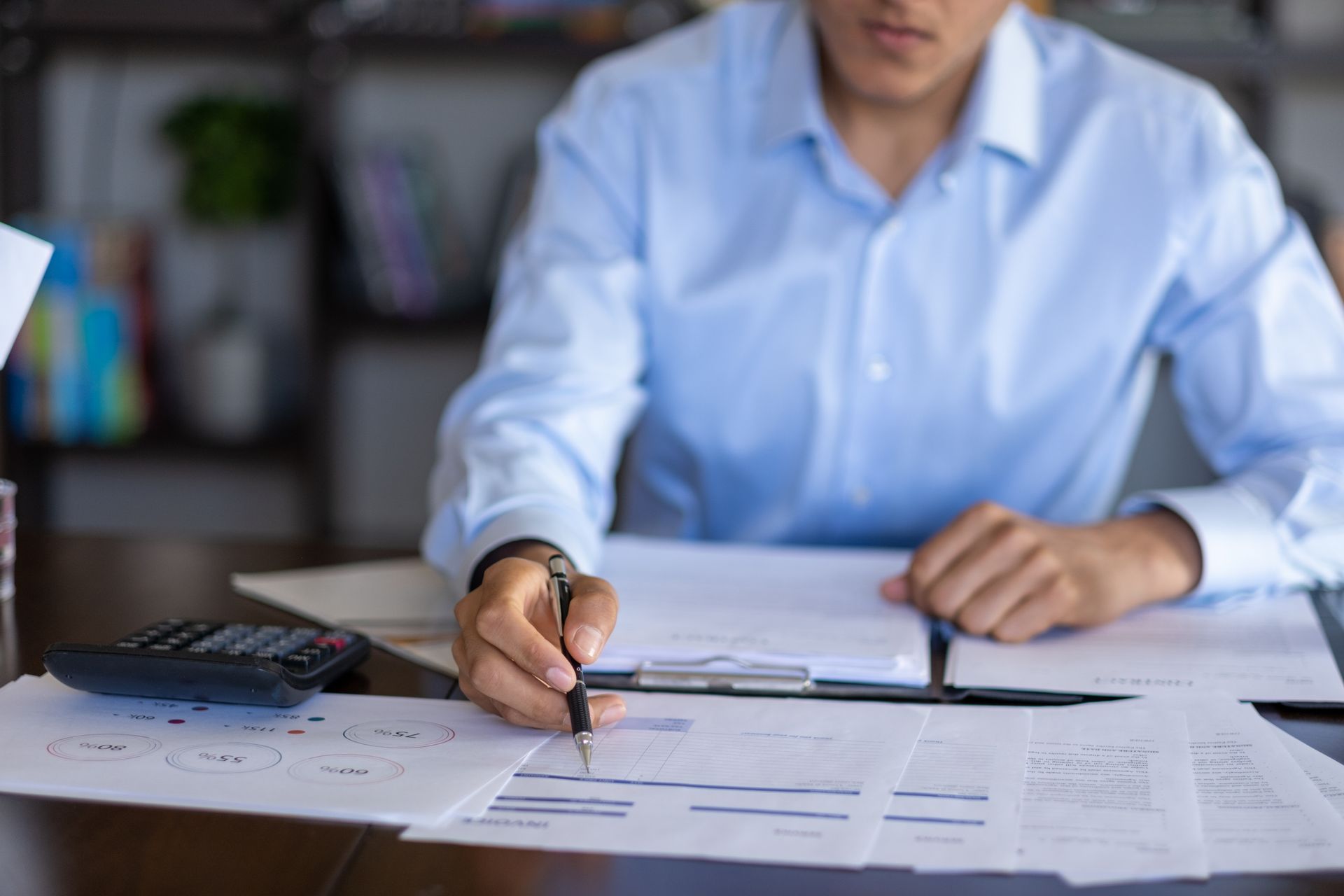 Una persona con una camicia azzurra punta la penna verso documenti finanziari vicino a una calcolatrice su una scrivania.