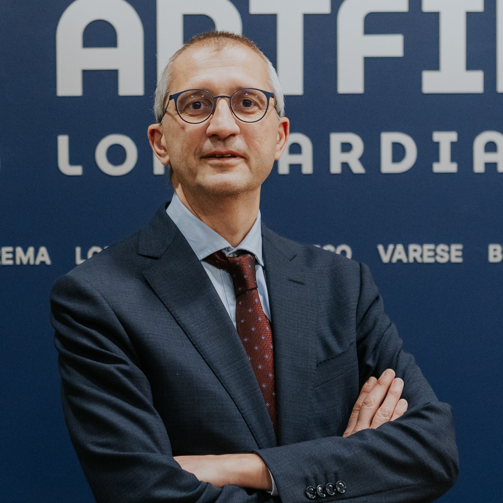 Uomo con gli occhiali, abito, cravatta rossa, sorridente; sfondo rosso.