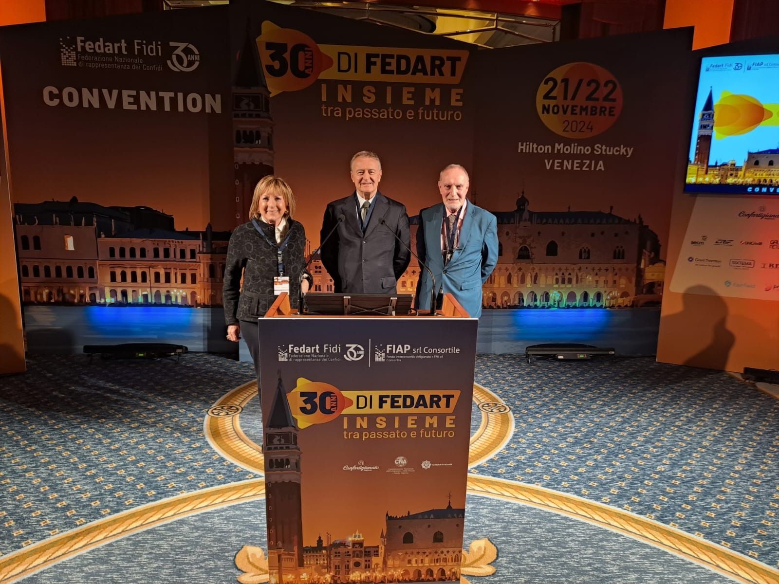 Tre persone in piedi dietro un podio durante una convention a Venezia. Sul podio è scritto