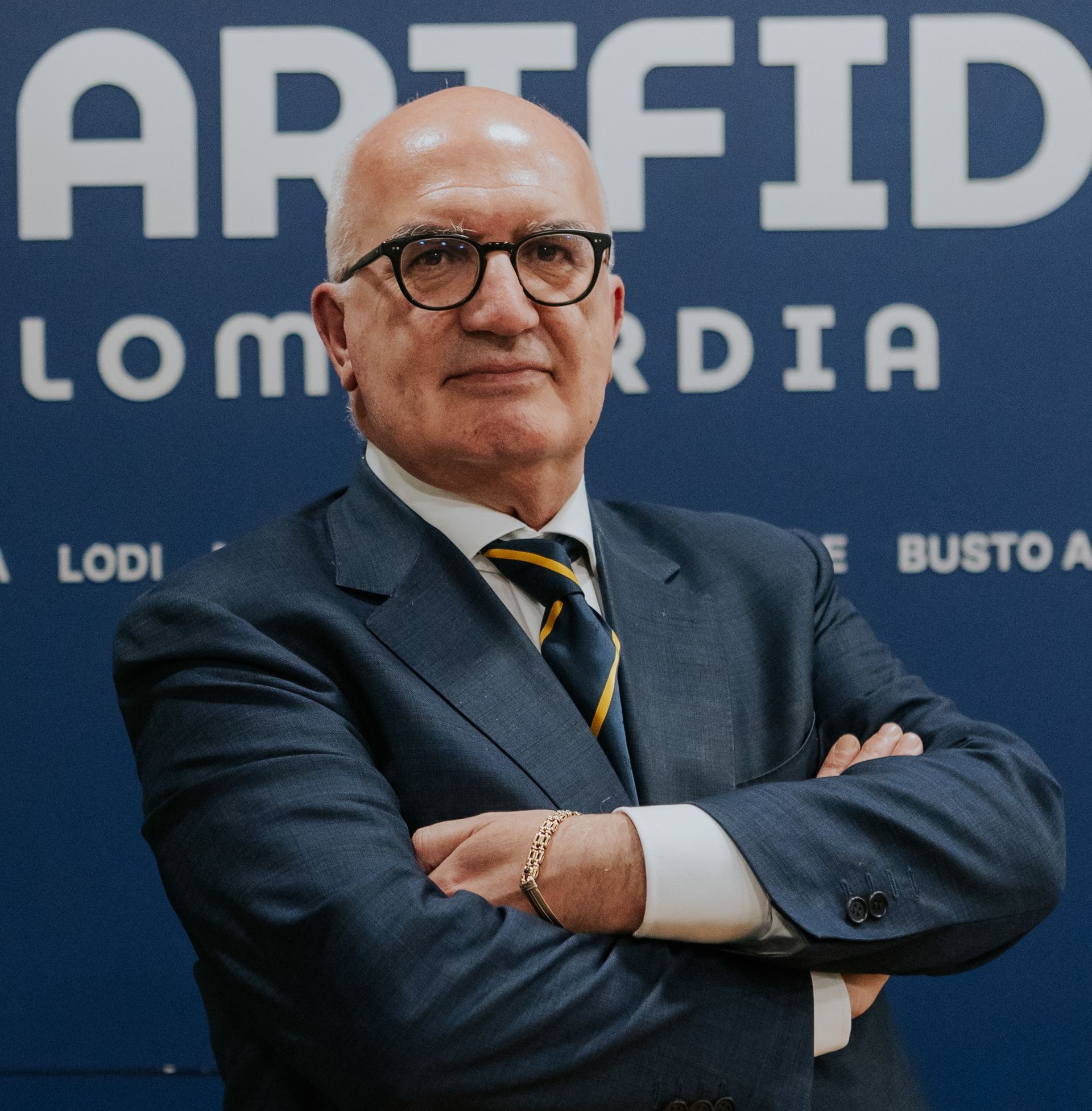 Uomo con occhiali e barba, in abito su sfondo rosso, sorridente.