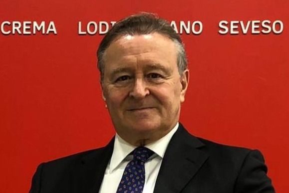 Uomo in giacca e cravatta su sfondo rosso, con un leggero sorriso. La scritta sopra recita 