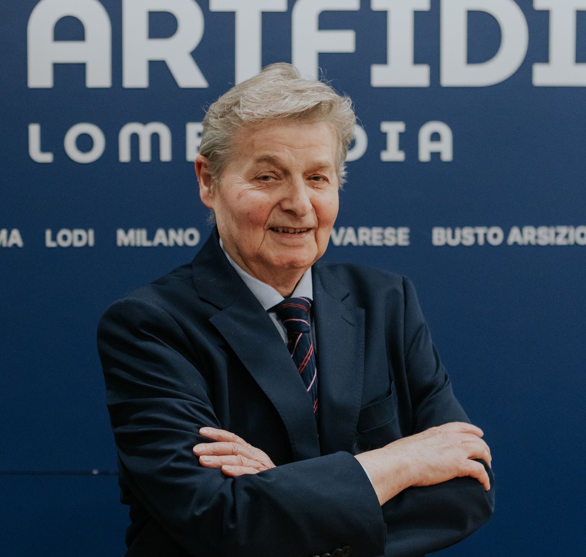 Uomo in giacca e cravatta su sfondo rosso; guarda direttamente l'osservatore.