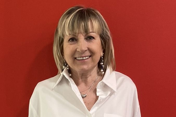 Donna con i capelli biondi che indossa una camicia bianca e sorride su uno sfondo rosso.