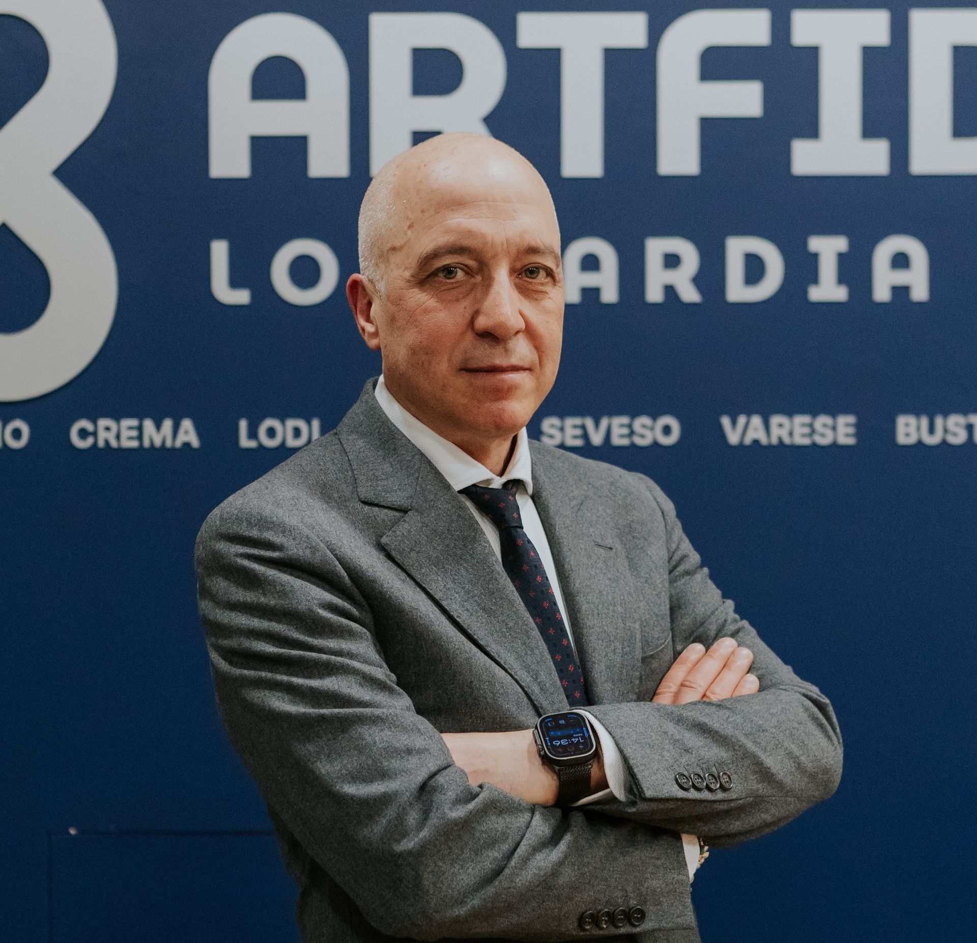 Uomo calvo in giacca e cravatta su sfondo rosso, sorridente verso la telecamera.