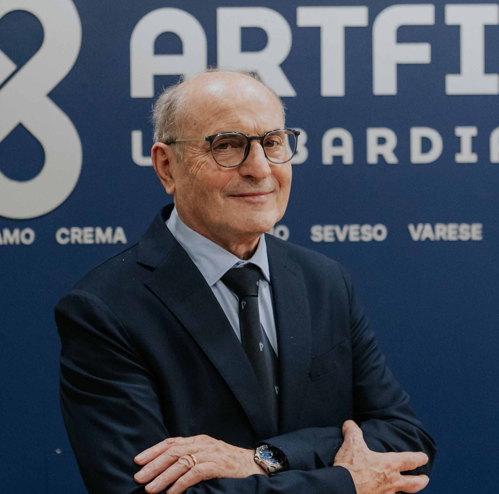 Uomo con gli occhiali, sorridente, che indossa un maglione e una cravatta, su sfondo rosso.