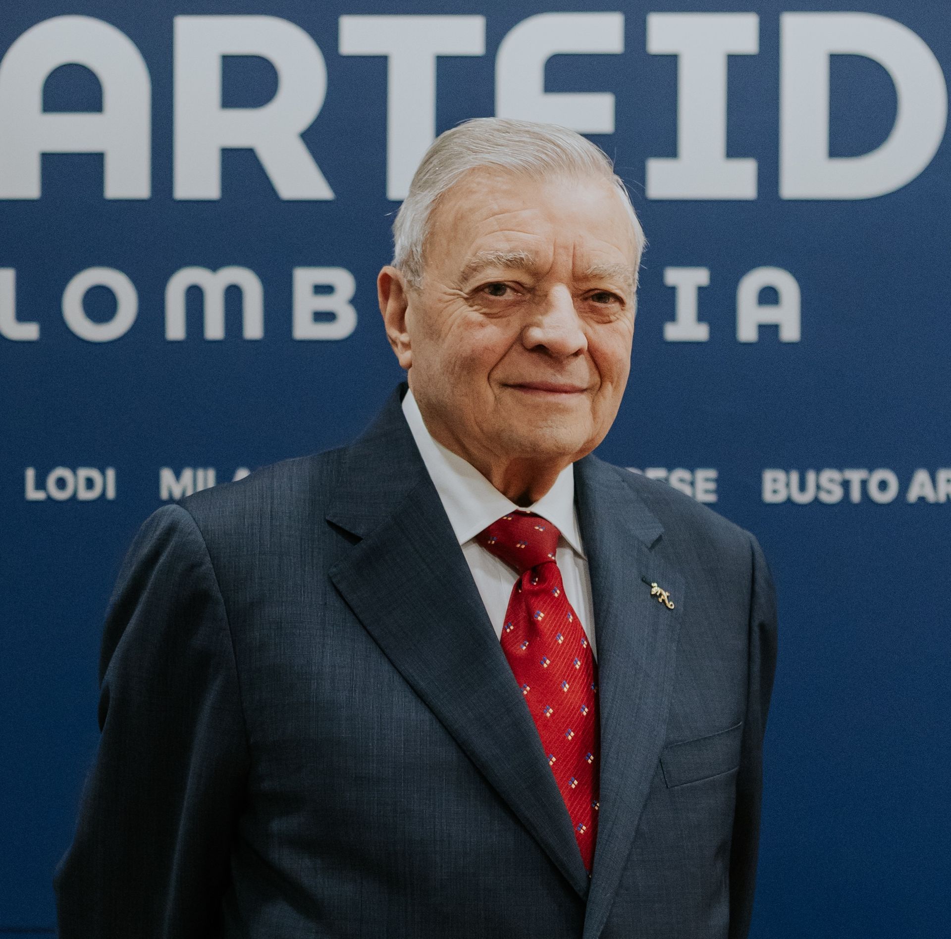 Un uomo in giacca e cravatta sorride, appoggiato a un muro rosso.