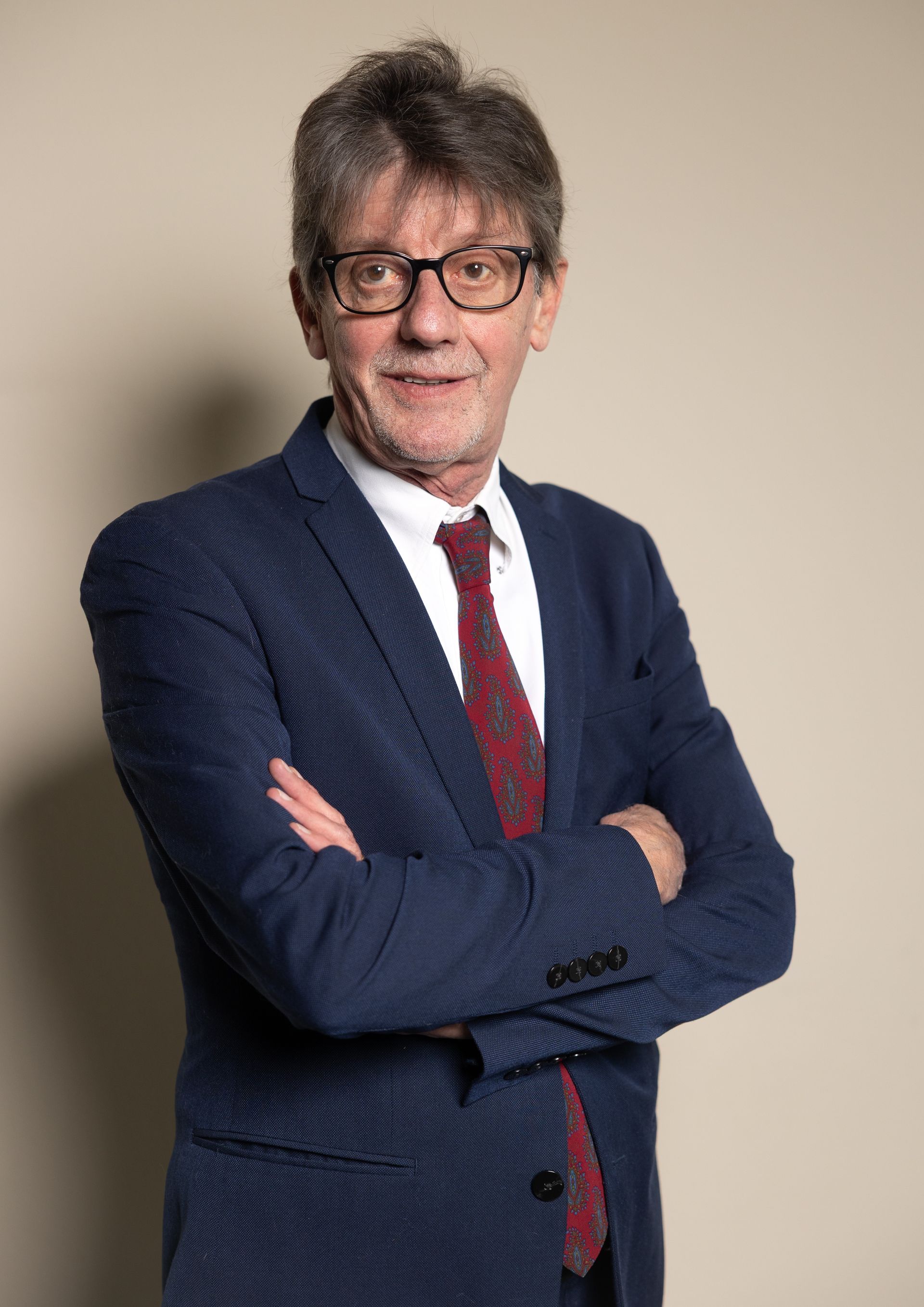 Un uomo in giacca e occhiali sorride, in piedi contro un muro rosso.