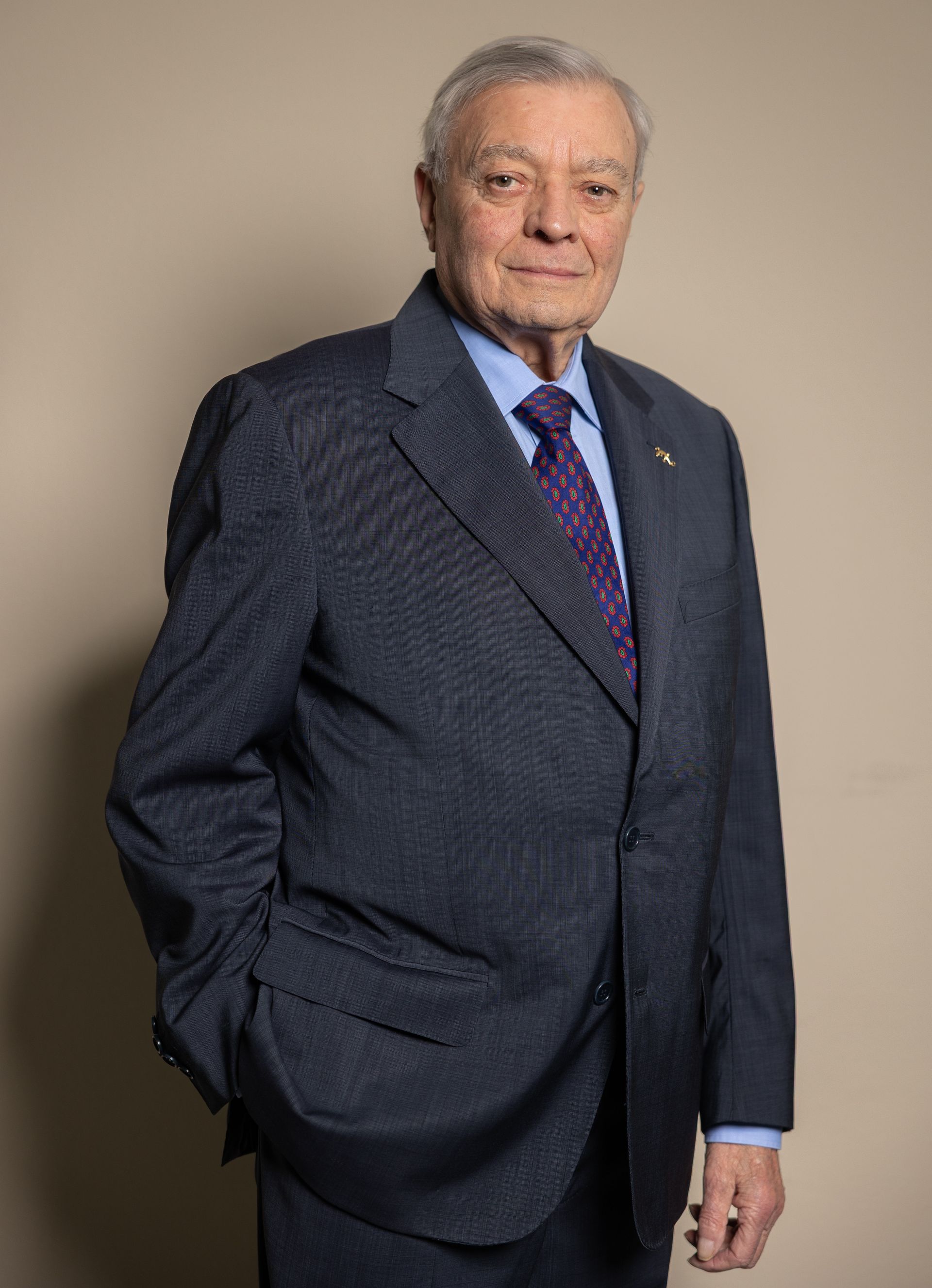 Un uomo in giacca e cravatta sorride, appoggiato a un muro rosso.