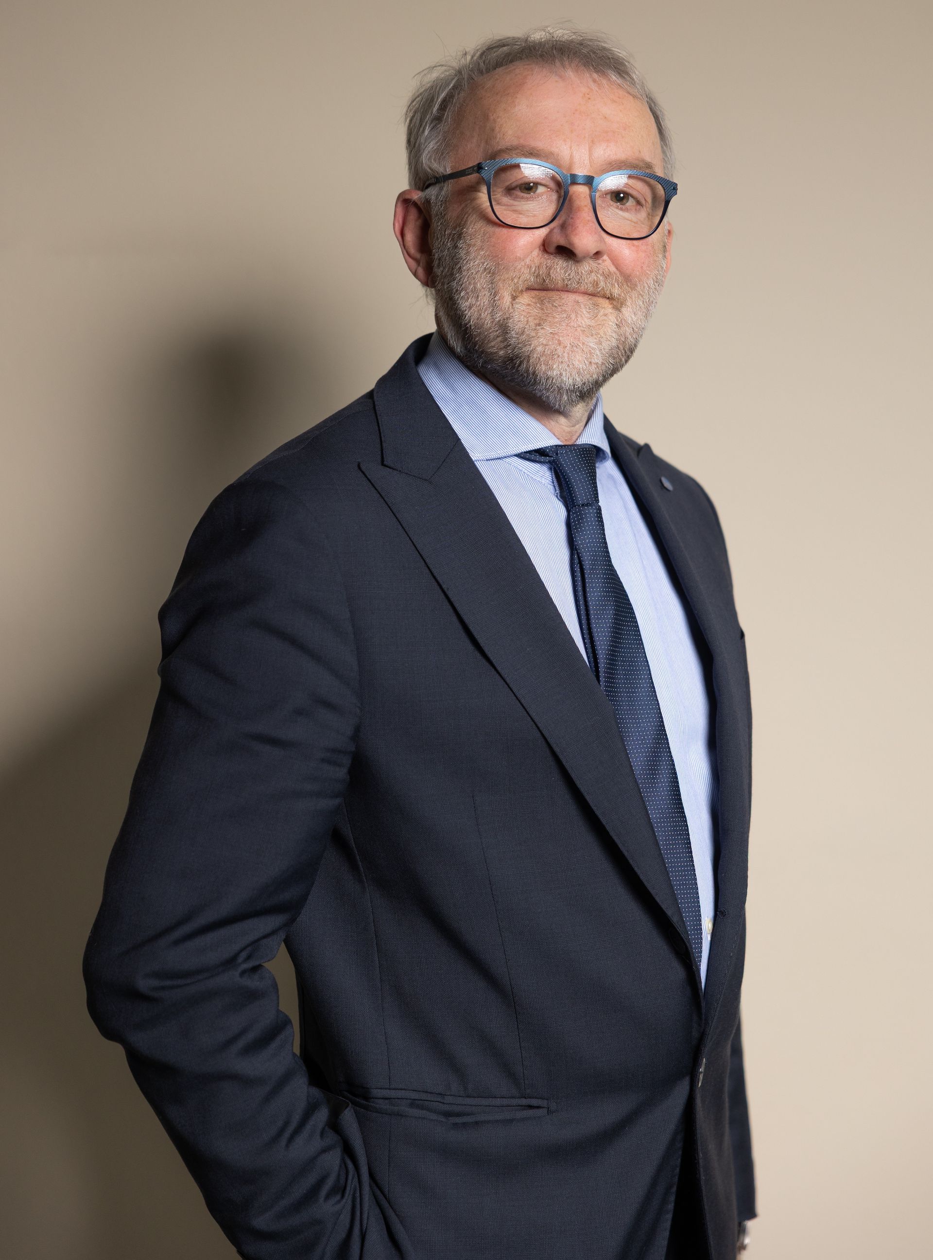 Uomo con occhiali e barba che indossa un abito, su uno sfondo rosso.