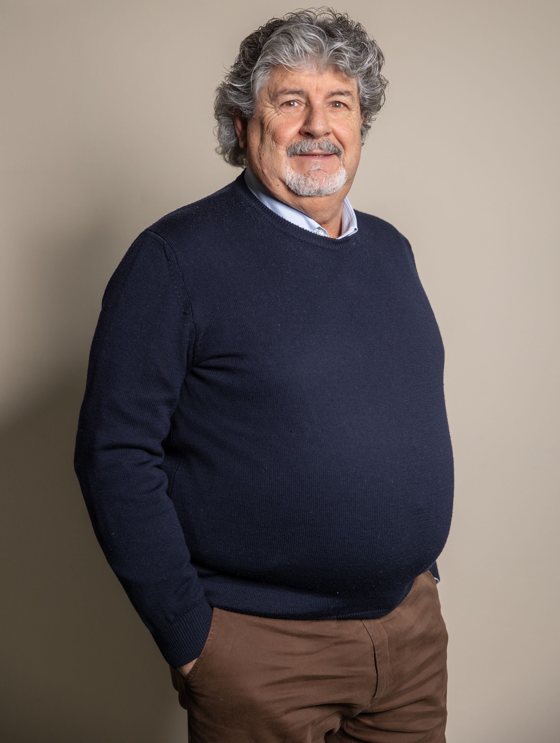 Uomo con barba e capelli grigi, vestito in giacca e cravatta, sorridente su sfondo rosso.