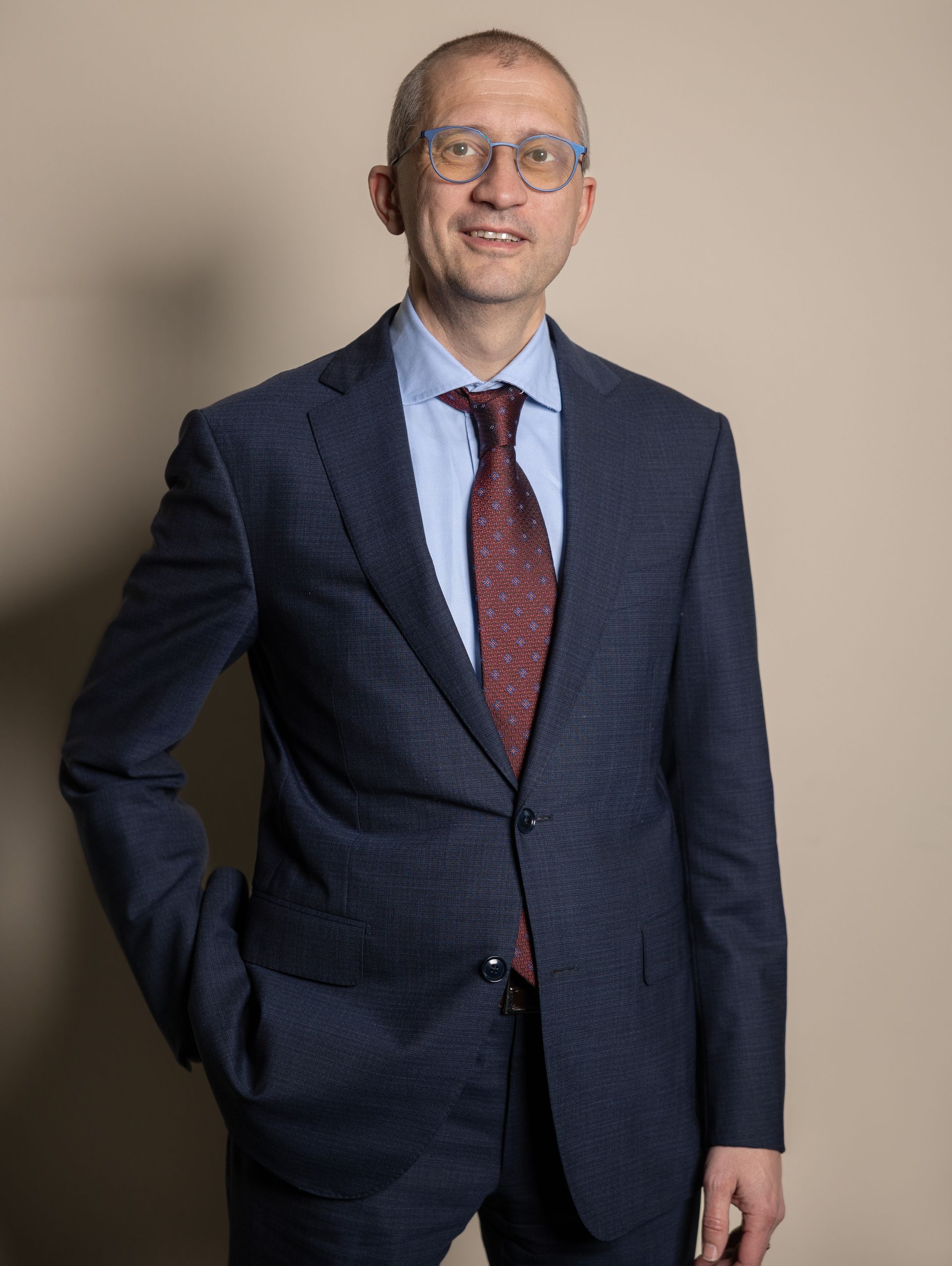 Uomo con gli occhiali, abito, cravatta rossa, sorridente; sfondo rosso.