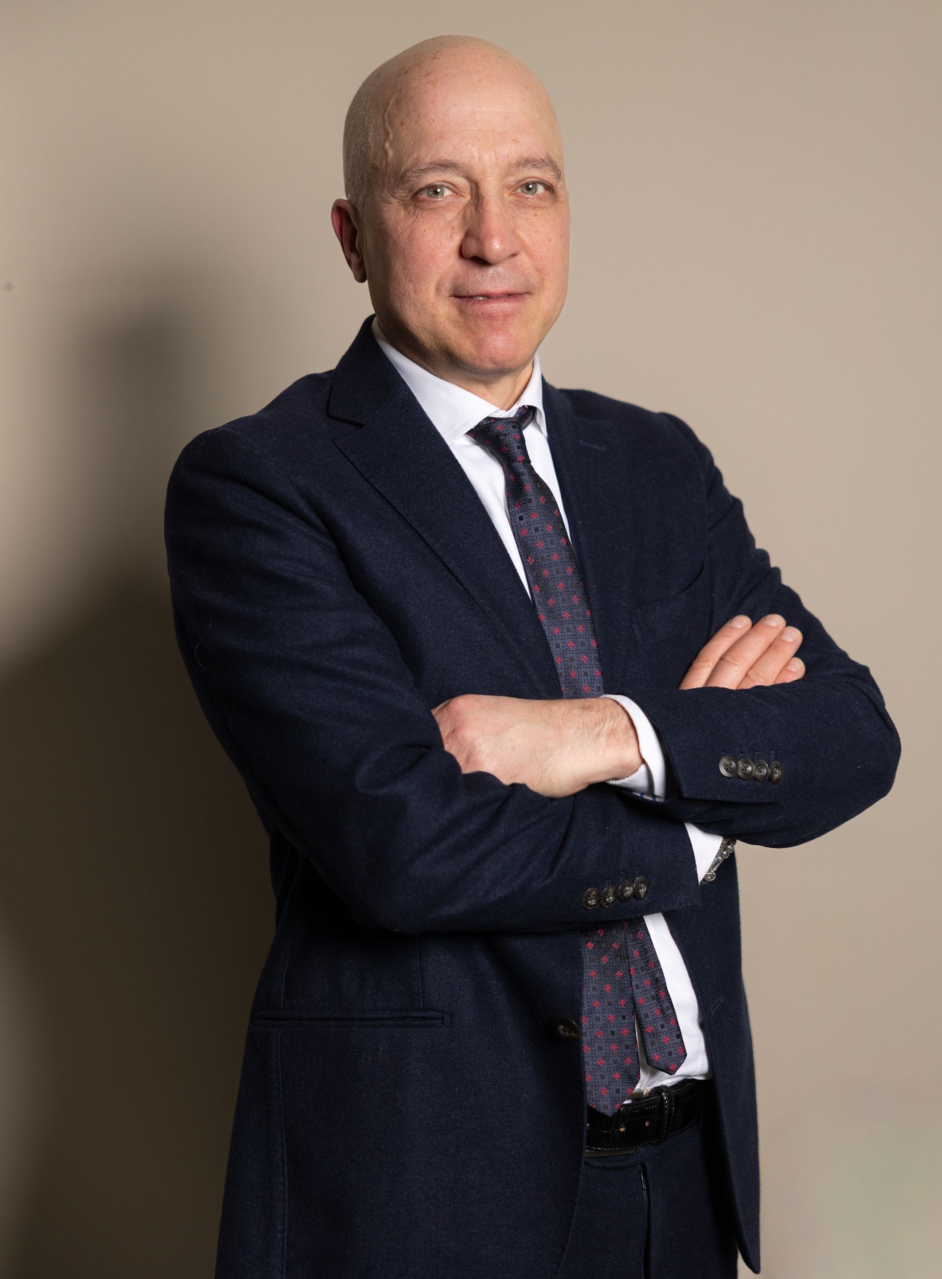 Uomo calvo in giacca e cravatta su sfondo rosso, sorridente verso la telecamera.