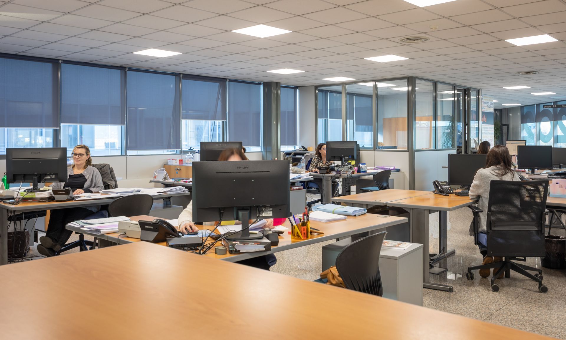 Tre impiegati seduti alla scrivania, uno sorridente, giacca blu, computer portatile, monitor, ufficio buio.