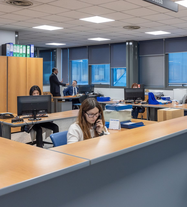 Tre impiegati seduti alla scrivania, uno sorridente, giacca blu, computer portatile, monitor, ufficio buio.