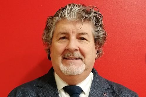 Uomo con barba e capelli grigi, vestito in giacca e cravatta, sorridente su sfondo rosso.