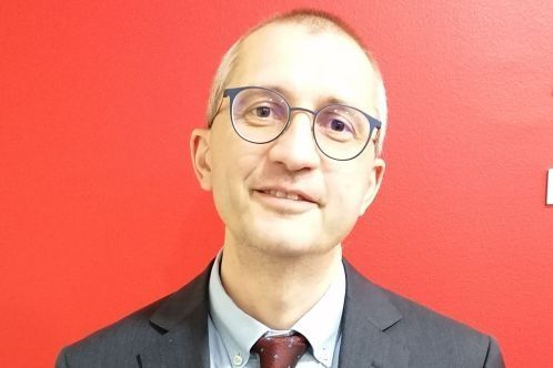 Uomo con gli occhiali, abito, cravatta rossa, sorridente; sfondo rosso.