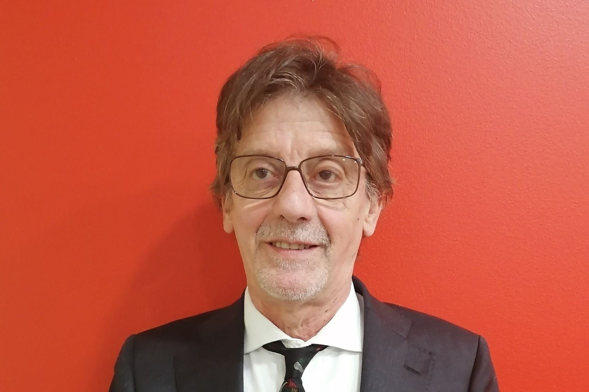 Un uomo in giacca e occhiali sorride, in piedi contro un muro rosso.