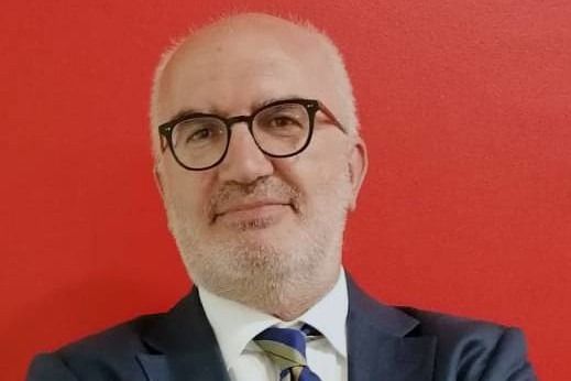 Uomo con occhiali e barba, in abito su sfondo rosso, sorridente.