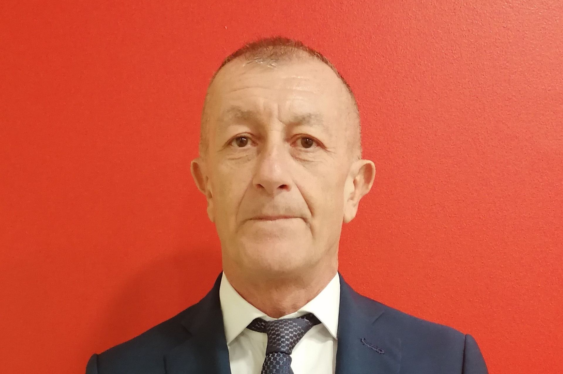 Uomo in giacca e cravatta su sfondo rosso, che guarda avanti.