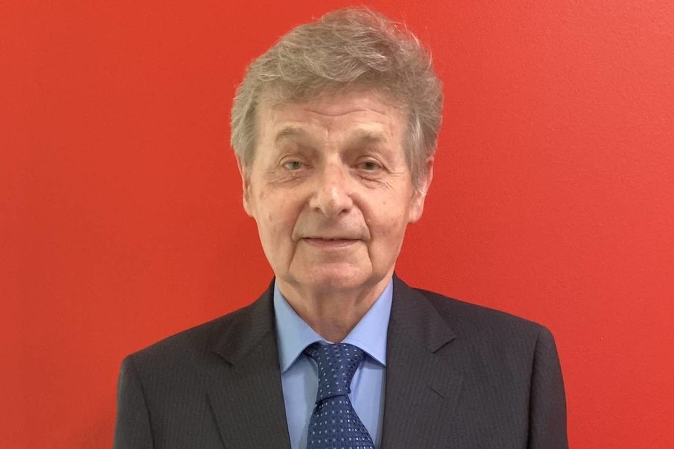 Uomo in giacca e cravatta su sfondo rosso; guarda direttamente l'osservatore.