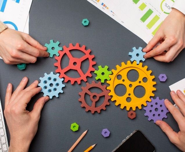 Mani che dispongono ingranaggi colorati su una superficie scura, con grafici e una tastiera nelle vicinanze.