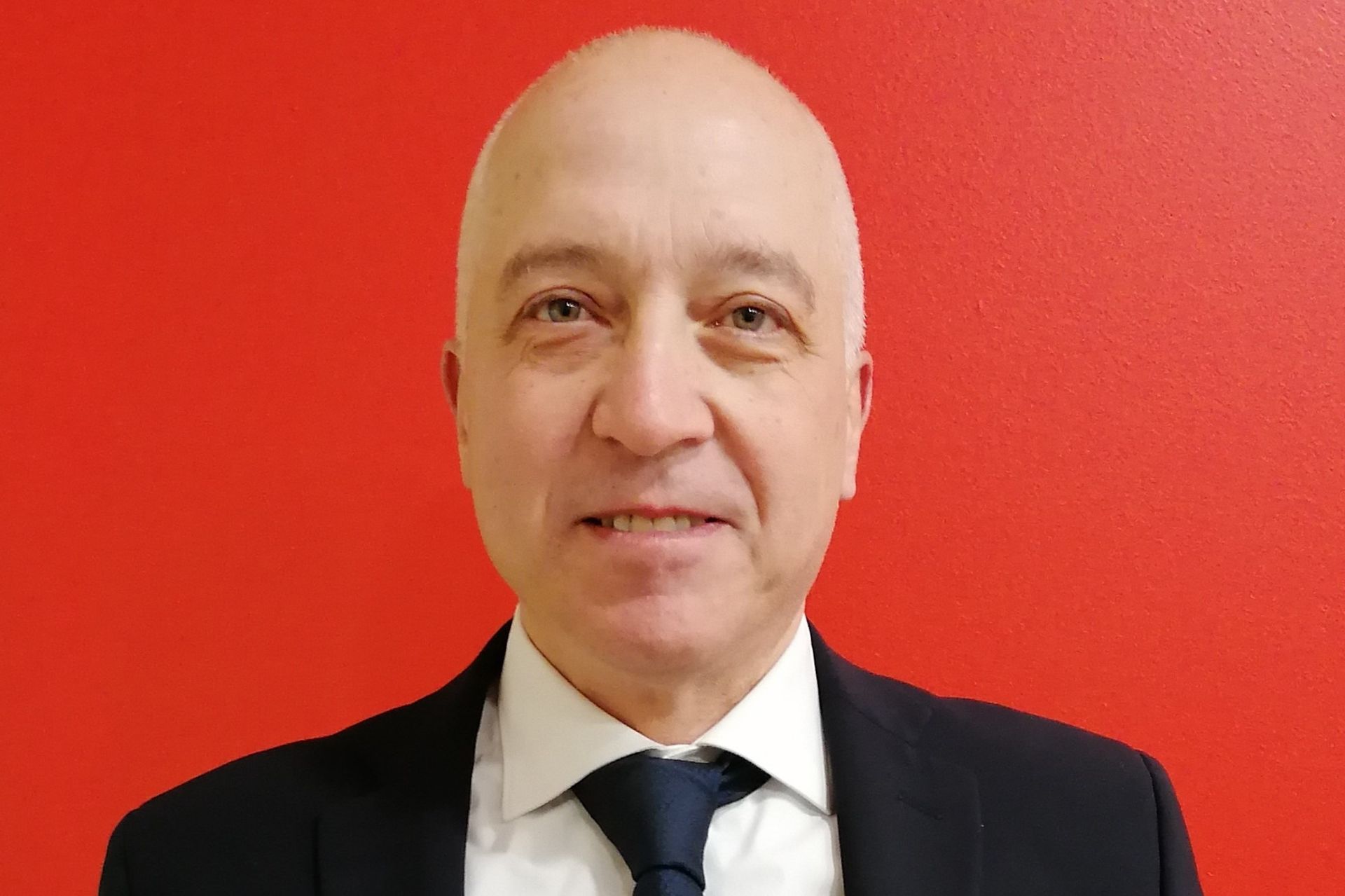 Uomo calvo in giacca e cravatta su sfondo rosso, sorridente verso la telecamera.