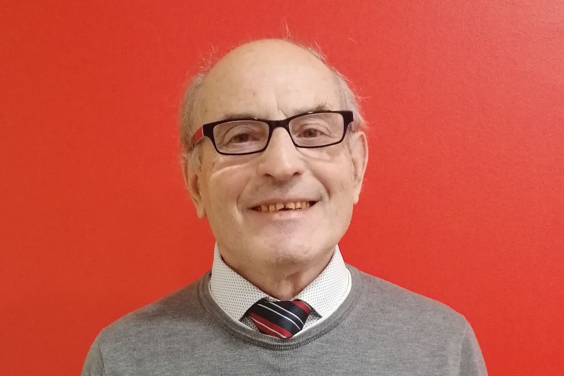 Uomo con gli occhiali, sorridente, che indossa un maglione e una cravatta, su sfondo rosso.