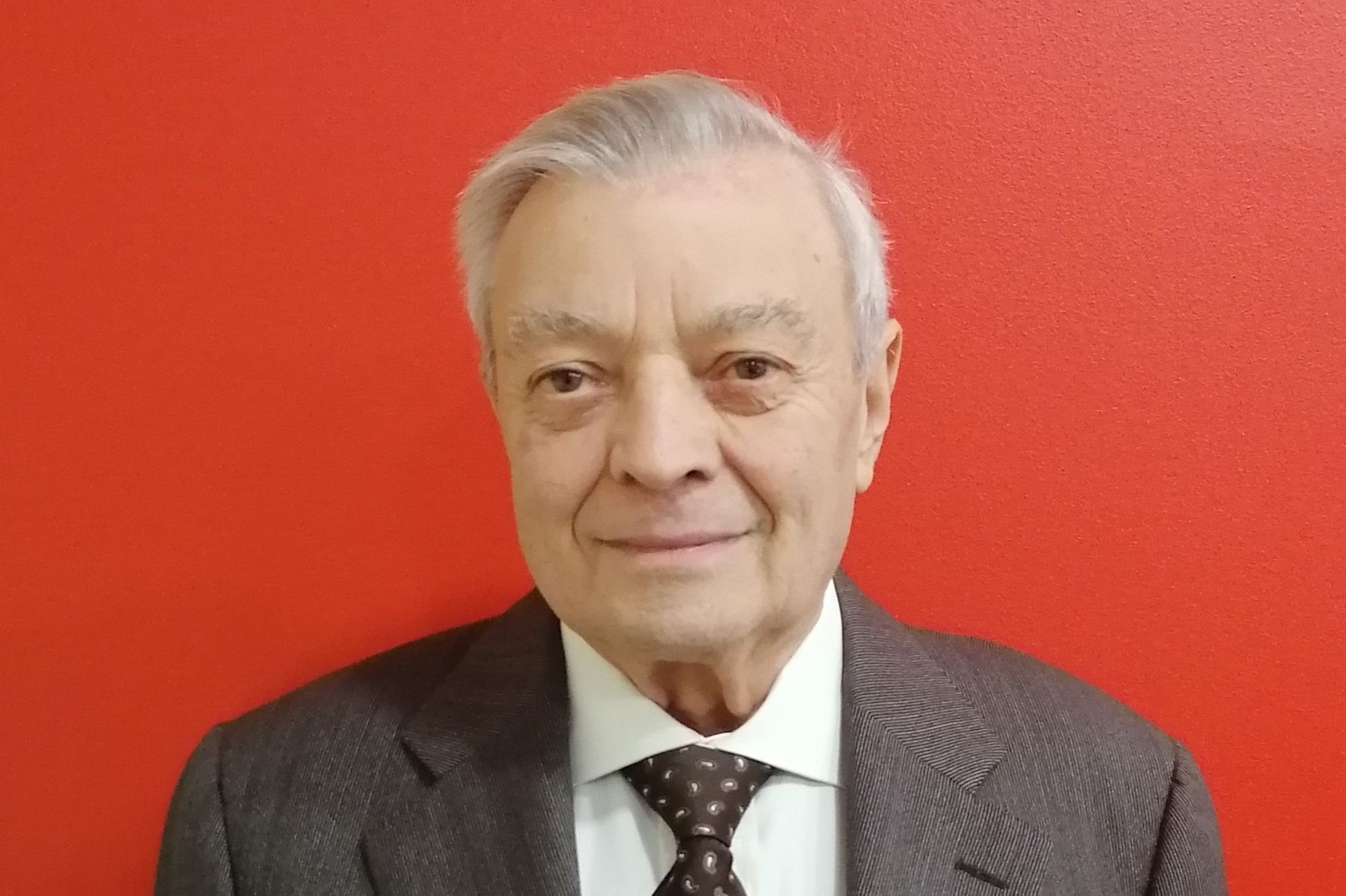 Un uomo in giacca e cravatta sorride, appoggiato a un muro rosso.