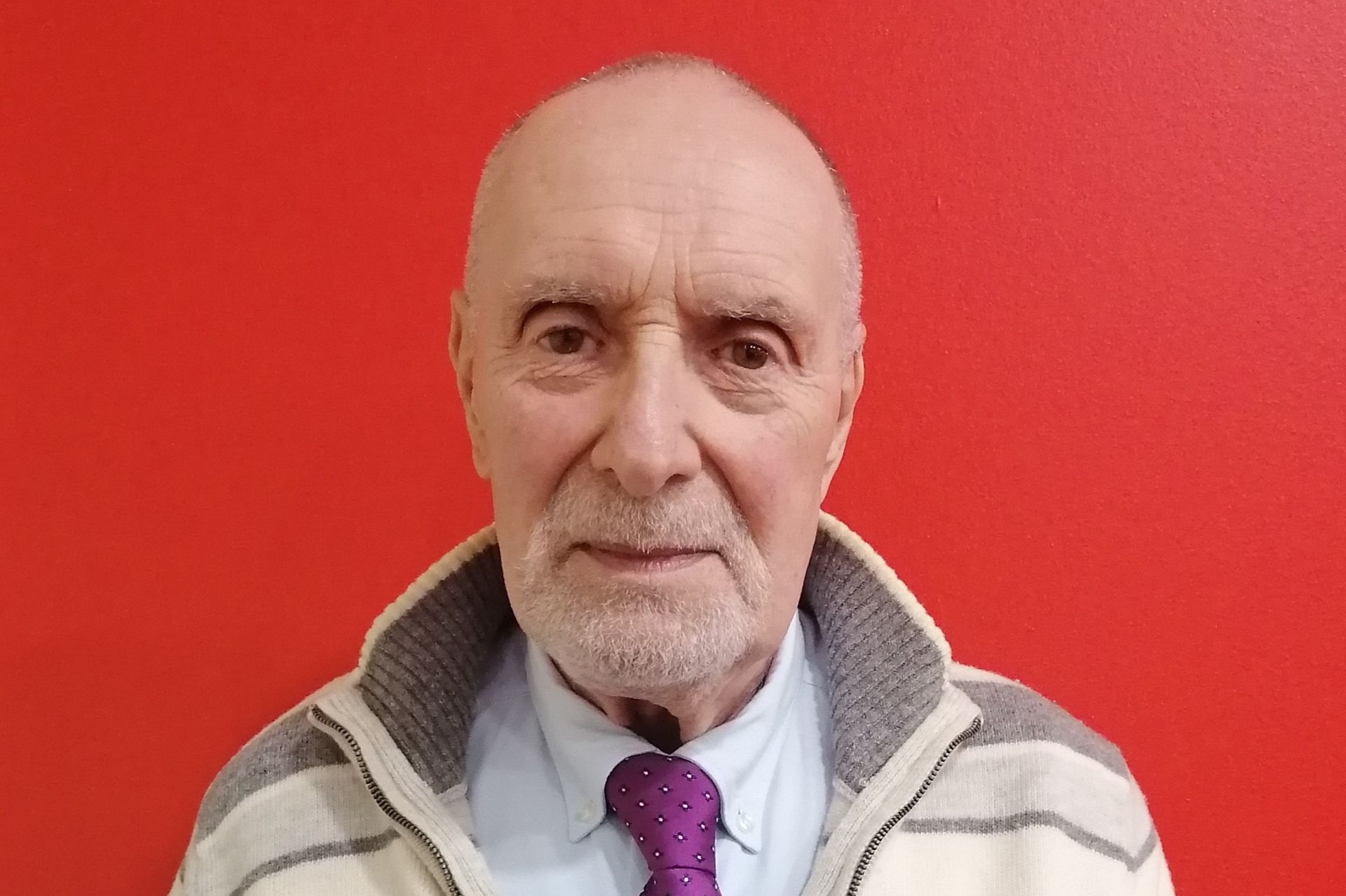 Uomo calvo con barba, maglione e cravatta, su sfondo rosso.