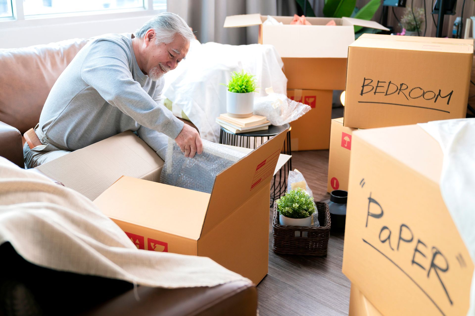 An elderly man is sitting on a couch opening a cardboard box.