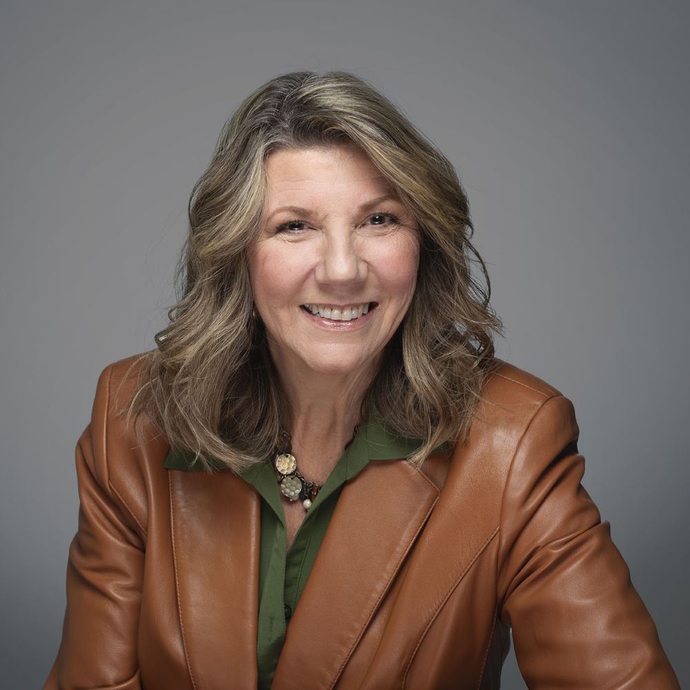 Woman with brown leather jacket and green shirt smiling in front of a gray backdrop.