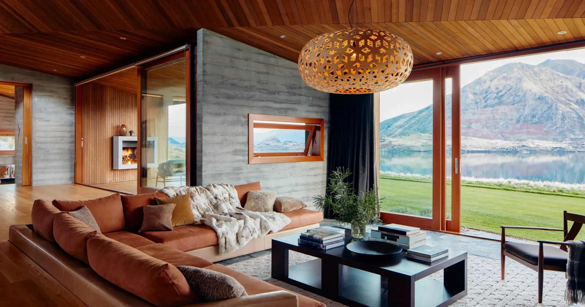 Modern living room with orange sectional, wooden ceiling, and mountain view through large doors.