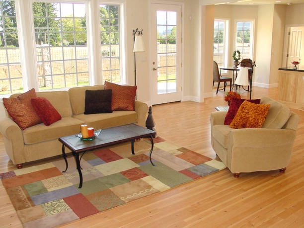Living room with beige couch, patterned rug, large windows, and dining area in the background.