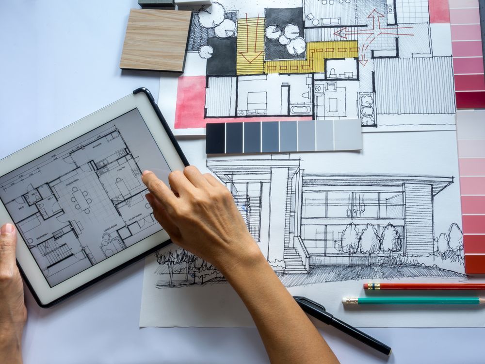 Person's hand using a tablet to review house plans, next to sketches, color swatches, and pencils on a white surface.