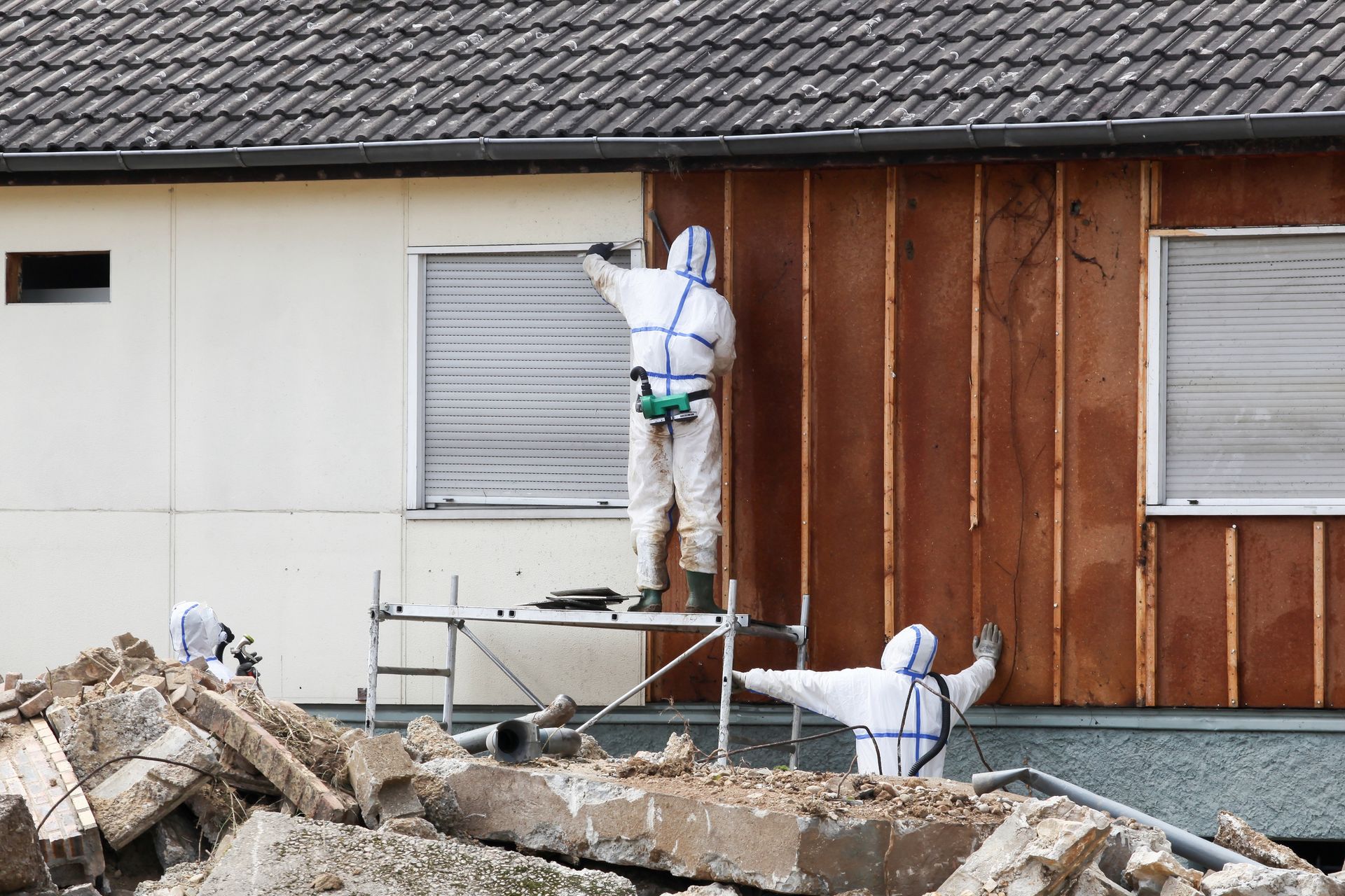 Workers in hazmat suits remove siding from a house with demolition debris in foreground.