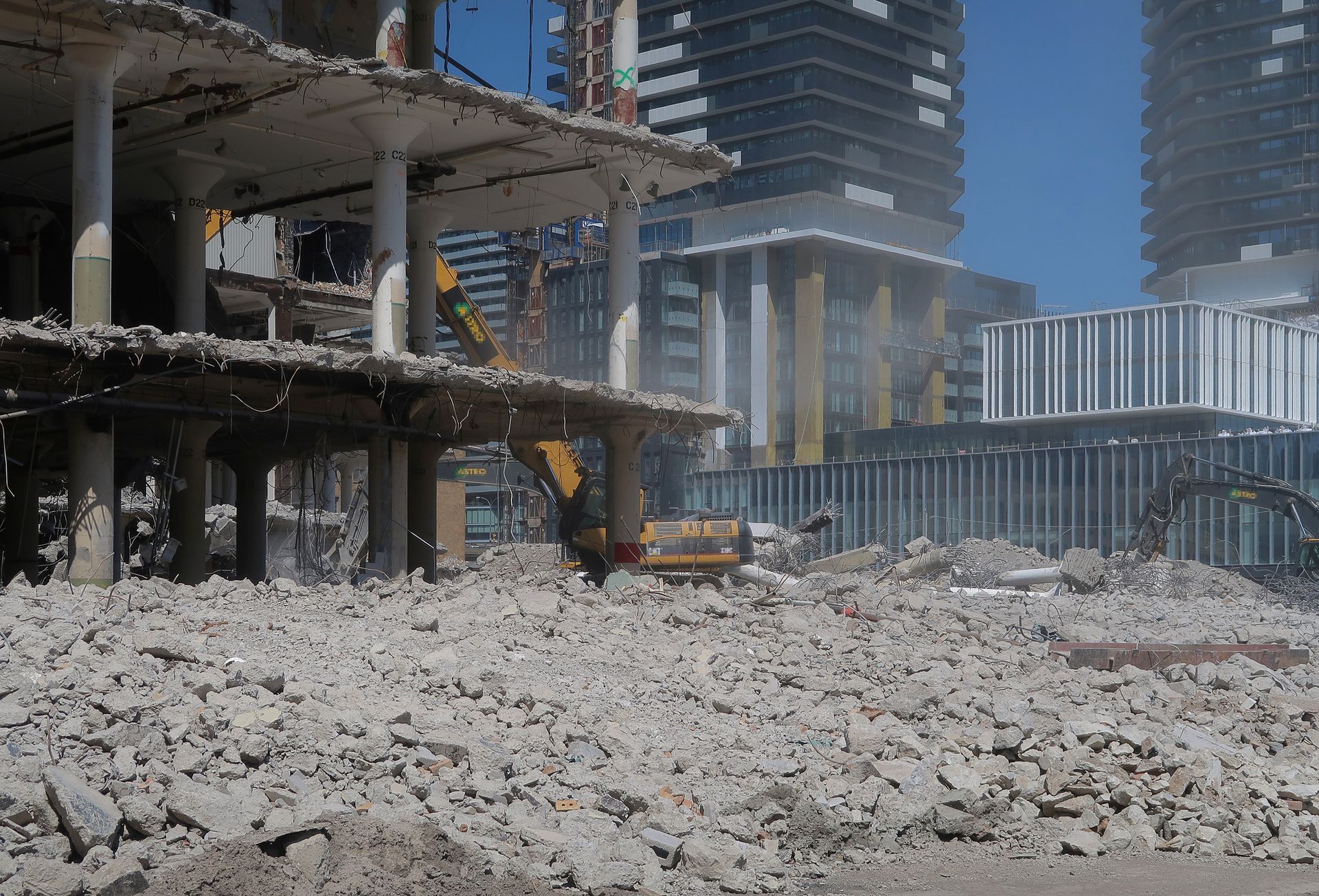 Building demolition with machinery, debris pile in foreground, modern city buildings in background.