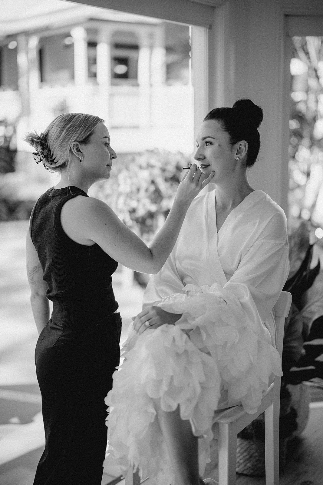 A woman is applying makeup to a bride in a black and white photo.