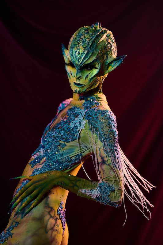 A woman with her body painted like a lizard is standing in front of a red curtain.