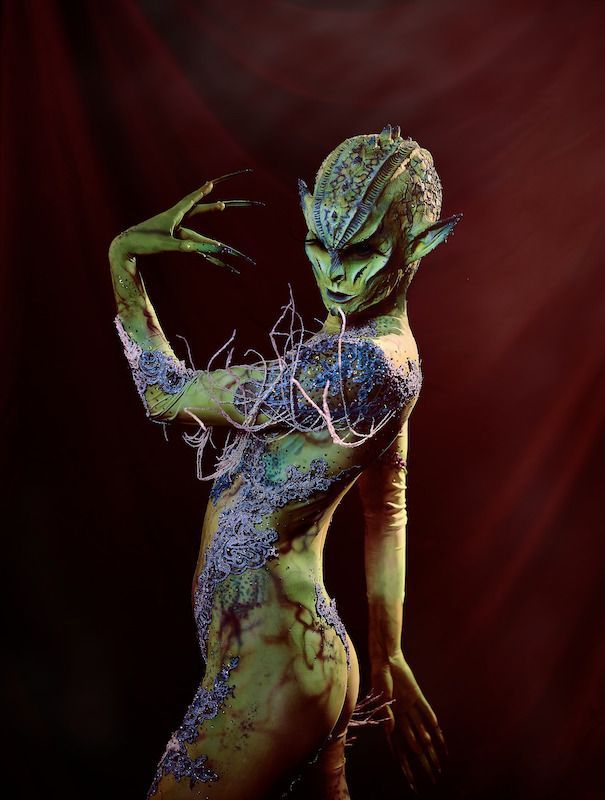 A woman with green body paint is standing in front of a dark background.